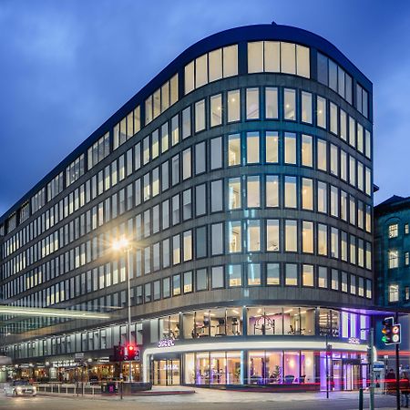Yotel Glasgow Esterno foto The photo shows a modern office building with a distinctive curved shape and a facade made of large glass windows. It is illuminated against a twilight sky, giving it a contemporary appearance. The ground floor features large glass entrance areas, li