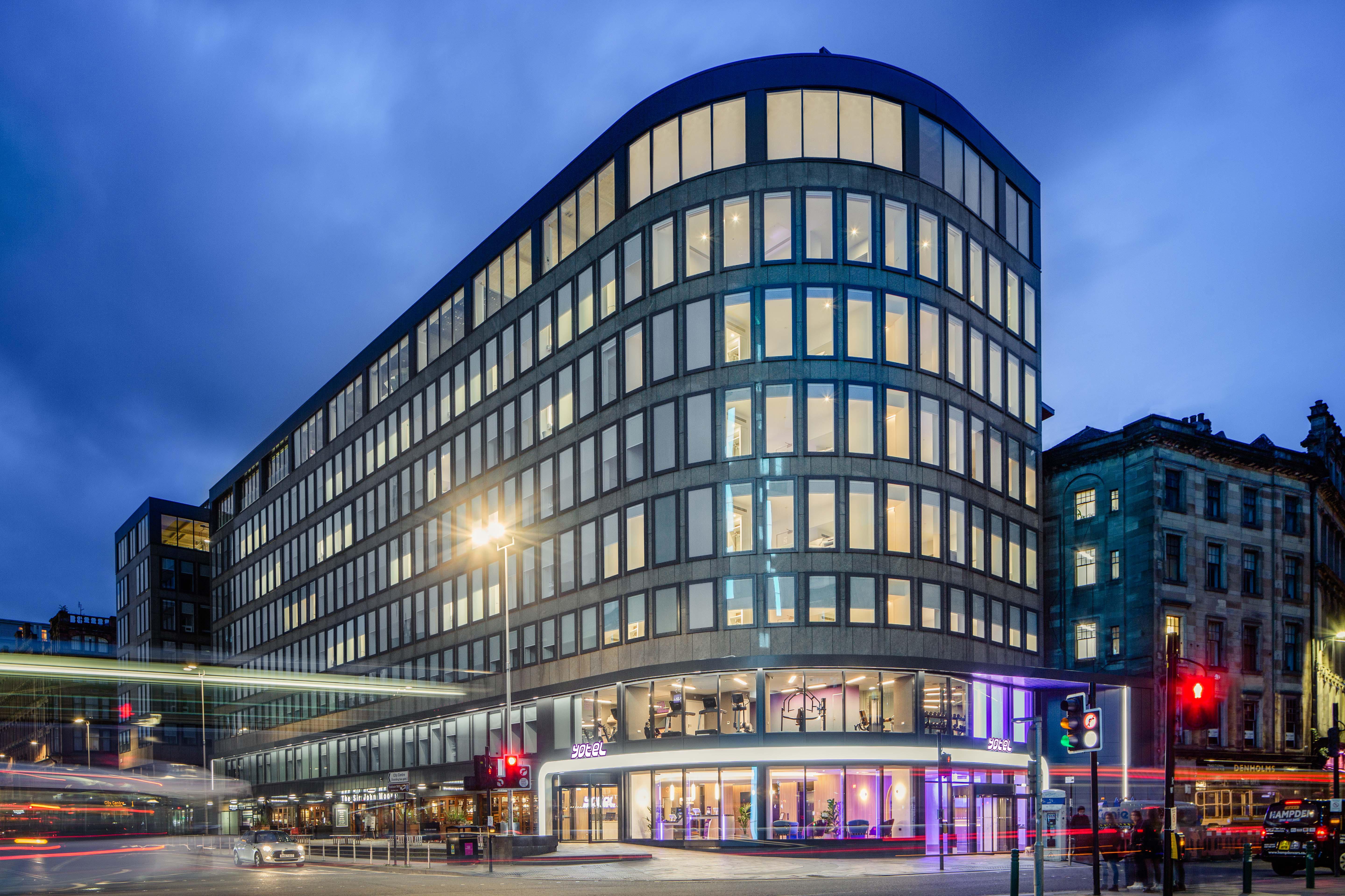 Yotel Glasgow Esterno foto The photo shows a modern office building with a distinctive curved shape and a facade made of large glass windows. It is illuminated against a twilight sky, giving it a contemporary appearance. The ground floor features large glass entrance areas, li